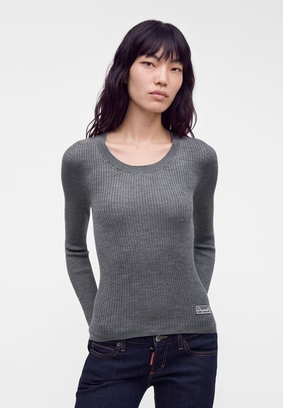 Femme aux longs cheveux foncés portant un pull gris côtelé ajusté et un jean bleu foncé, debout devant un fond clair uni.