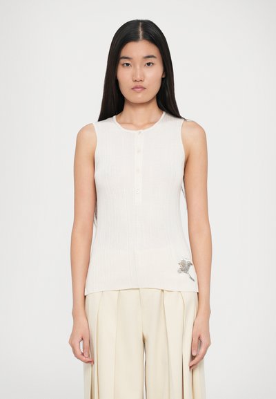 Débardeur côtelé crème avec encolure ronde, cinq boutons et un détail floral brodé en bas à gauche. Associé à un pantalon beige.