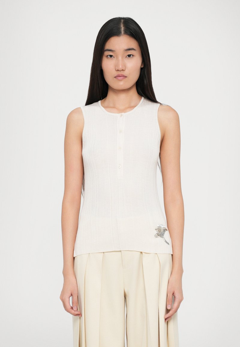 Kermarin raidallinen hihaton paita, jossa on pyöreä kaula-aukko, viisi nappia ja laidan vasemmalla puolella brodeerattu kukkakuva. Yhdistetty vaaleanbeigeihin housuihin.