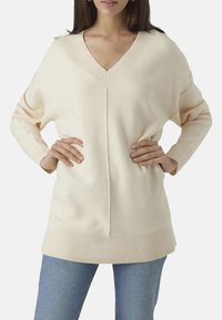 Pull V beige clair à col en V avec des manches longues, des poignets et un ourlet côtelés, présentant un détail de couture central et une coupe décontractée.