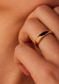 Gouden ring met een gepolijste oppervlakte, voorzien van een gladde, gebogen vorm. Draagbaar aan een hand met zichtbare huidtextuur en subtiele schaduwen.