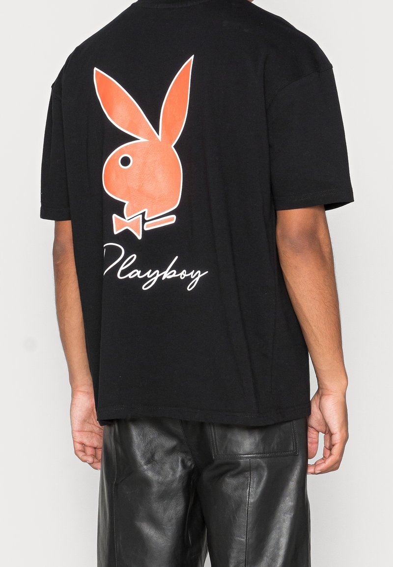 Personne portant un t-shirt noir avec un grand logo lapin Playboy orange et un texte blanc dans le dos, associé à un pantalon en cuir noir.
