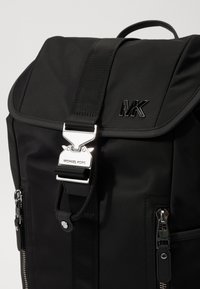 Black nylon backpack features a fold-over flap, silver hardware, 'MK' logo, and zippered pockets. Smooth texture with adjustable straps.