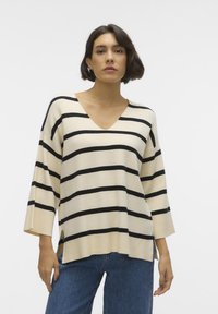 Pullover beige con strisce orizzontali nere, scollo a V, maniche ampie, spacchi laterali e una consistenza morbida. Indossato con jeans blu.