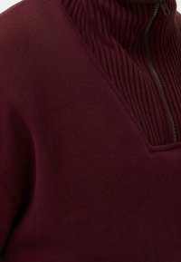 Sudadera burdeos con cuello alto que presenta una textura acanalada. Tiene un cierre de cremallera y tela lisa en el cuerpo.