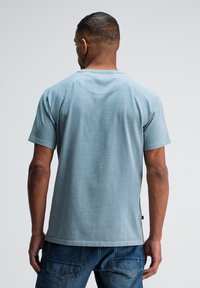 Man in een lichtblauw kortemouwen shirt en donkerblauwe jeans, staand met de rug naar de camera tegen een effen grijze achtergrond.