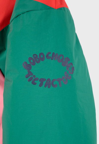 Veste colorée avec un corps vert, des accents roses et rouges, en tissu doux, et un logo graphique bleu audacieux sur le devant.