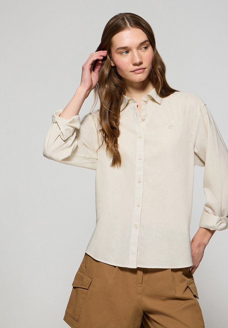 Beżowa koszula zapinana na guziki wykonana z lekkiego materiału, z kołnierzykiem i podwiniętymi rękawami, z subtelnym logo na piersi. W zestawie z khaki szortami cargo.