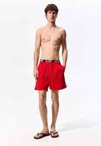 Costume da bagno rosso con vita elastica nera, dotato di cordoncino e tasche laterali. Il modello è in piedi a piedi nudi, indossando ciabatte nere.