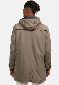 Vīrietis ar tumšiem dreads, ģērbies garā pelēkā mētelī ar kapuci un melnām regulējamām piedurknēm, redzams no aizmugures.