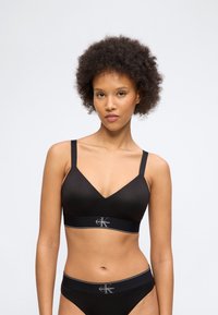 Γυναίκα με σγουρά μαλλιά, φορώντας μαύρο bralette Calvin Klein και ταιριαστό εσώρουχο, στέκεται μπροστά σε ένα απλό ανοιχτό φόντο.