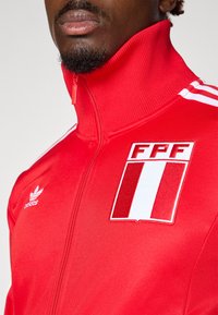 Punane spordijakk kõrge kraega, millel on valged triibud õlgadel ja suure FPF logo kilp. Valmistatud siledast, venitavast kangast.