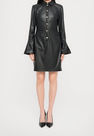 Femme portant une robe en cuir noire à manches longues boutonnée avec des poignets évasés et des escarpins noirs à bouts pointus, debout devant un fond uni.