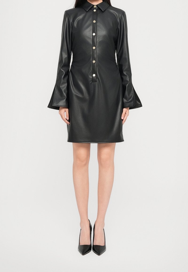 Femme portant une robe en cuir noire à manches longues boutonnée avec des poignets évasés et des escarpins noirs à bouts pointus, debout devant un fond uni.
