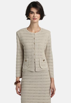 Kobieta ubrana w beżową, teksturowaną dzianinaną kurtkę z guzikami i pasującą spódnicę, uzupełniona złotymi kolczykami i brązową bransoletką.