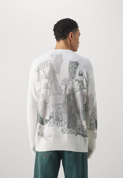 Weißer Strickpullover mit einem kunstvollen grauen Druck auf dem Rücken, versehen mit Text und visuellen Elementen, sowie einem gerippten Saum. Kombiniert mit einer Jeans.