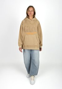 Beige Oversized-Hoodie mit Fronttasche, strukturiertem "GRC"-Logo in erhabenem Design und orangefarbener Schrift. Kombiniert mit hellblauen weit geschnittenen Jeans und weißen Sneakern.