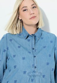 Camicia di jeans azzurro chiaro con motivi di fiocchi ricamati, chiusura con bottoni e colletto classico. Il tessuto ha un aspetto morbido e strutturato.