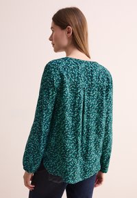 Blusa verde a fantasia con maniche lunghe arricciate, scollatura rotonda e vestibilità morbida. Presenta un motivo di foglie in diverse tonalità di verde.