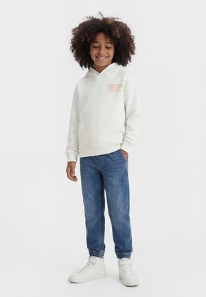 Enfant aux cheveux bouclés portant un sweat à capuche blanc, un jean bleu avec des poignets élastiques et des baskets blanches, debout et souriant avec une main dans la poche.