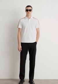 Balta polo marškinėliai su smėlio spalvos peties akcentais, trumpomis rankovėmis ir apykakle, derinami su juodomis kelnėmis ir tamsiais akiniais nuo saulės. Lygus audinio paviršius.