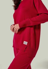 Roter Strickpullover mit V-Ausschnitt, gerippten Bündchen und seitlichen Schlitzen. Das Etikett unten links zeigt ein handschriftliches Logo.