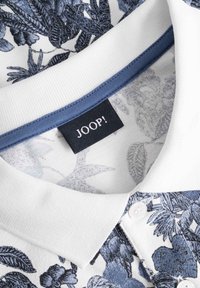 Vit polo skjorta med blått blommönster, krage och knäppning i enhetlig blå färg, med en liten svart etikett med "JOOP!" på insidan.