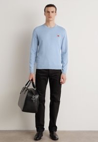 Hellesblaues gestricktes Sweatshirt, schwarze Hose, hält eine schwarze Lederhandtasche. Hat ein kleines Logo auf der Brust und eine glatte Textur.