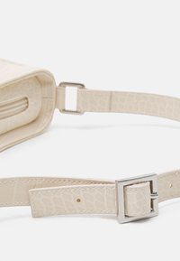 Sac à main crème en simili-crocodile avec une sangle à boucle en métal. Présente une texture lisse et un design structuré avec une fermeture à glissière.