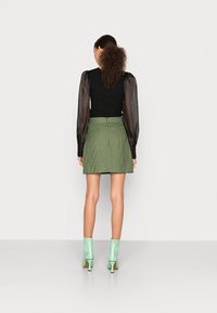 Une femme aux cheveux bouclés porte un haut noir et une jupe à carreaux verts, assortie de bottines mint green, tournée de dos contre un fond blanc.