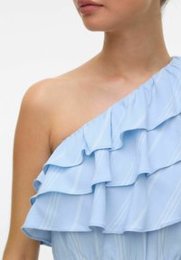 Robe bleue à une épaule avec des volants superposés à motifs rayés blancs, en tissu lisse, mettant en avant la texture et le design.