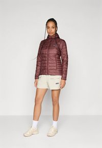 Veste isolante marron avec capuche, fermeture éclair et design matelassé. Associée à un short beige clair et des baskets beiges.
