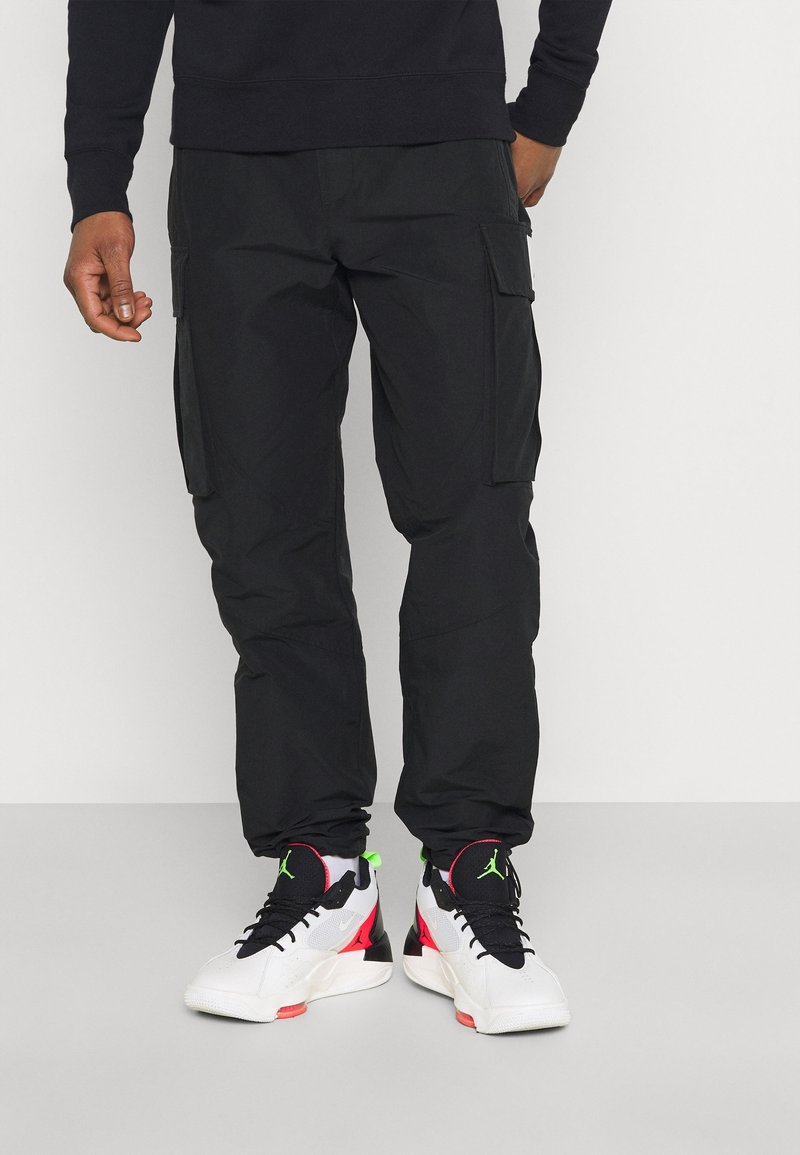 Personne portant un pantalon cargo noir et des baskets montantes blanches, noires et rouges avec des accents vert néon, debout sur un sol gris clair.