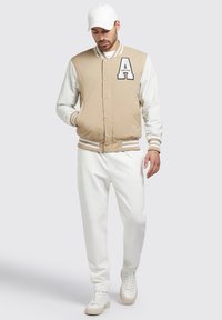 Jachetă bej de tip varsity cu mâneci crem, având un mare logo 'A' și manșete cu dungi. Purtați cu pantaloni albi și adidași.