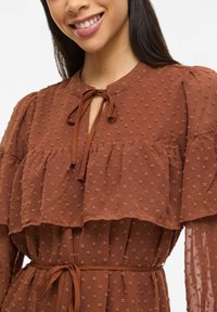 Robe marron avec un motif à pois texturé, des couches à volants, un col en trou de serrure avec lien, et une ceinture ajustée à la taille. Manches transparentes.