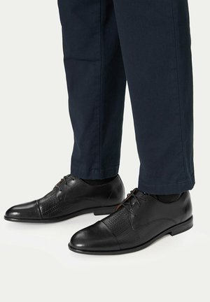 Schwarze Lederschuhe mit strukturierten Einsätzen, getragen zu dunkelblauen Hosen, vor einem schlichten hellen Hintergrund.