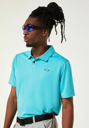 Polo turchese con colletto e maniche corte, caratterizzata da un logo nero sul petto a sinistra. L'individuo indossa occhiali da sole con lenti blu tinte.