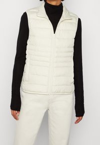 Hellbeige gesteppte Weste mit hohem Kragen, Frontreißverschluss und horizontalen Steppnähten. Über einem schwarzen Langarmshirt getragen. Glatte Textur.