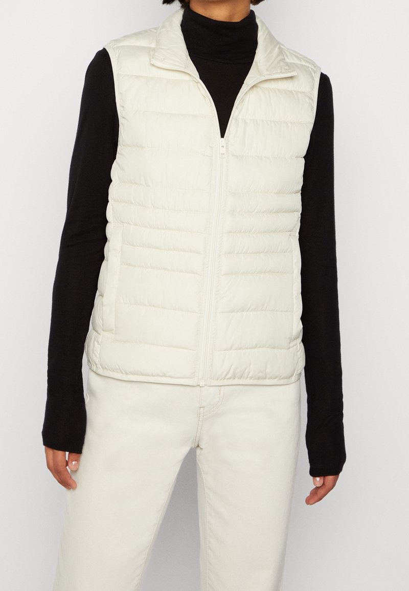 Hellbeige gesteppte Weste mit hohem Kragen, Frontreißverschluss und horizontalen Steppnähten. Über einem schwarzen Langarmshirt getragen. Glatte Textur.