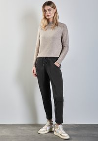 Suéter de punto beige con cuello alto, combinado con joggers negros que tienen bolsillos laterales y zapatillas de deporte de color canela y blanco. Textura suave en todo.