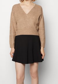 Pull en tricot beige à col en V, cropé, avec des manches longues, au-dessus d'une jupe évasée noire. Caractéristiques d'un tissu texturé et d'une taille cintrée.