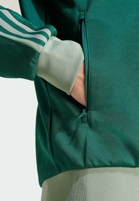 Grön sportjacka med strukturerat tyg, kontrasterande ljusgröna muddar och en zipficka på sidan.