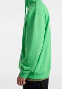 Osoba w jasnozielonym hoodie z troczkami, stojąca bokiem z widoczną ręką, w czarnych spodniach i oliwkowej koszuli pod spodem.