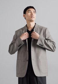 Hellgrauer Blazer mit Reverskragen, zwei Vordertaschen und schwarzen Knöpfen. Über einem schwarzen Hemd getragen, vor einem neutralen Hintergrund präsentiert.