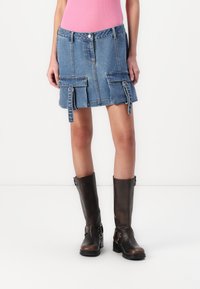 Farmer mini szoknya elülső zsebekkel, világoskék színben, nyers szegéllyel, állítható oldalszíjakkal, barna térd fölé érő csizmával párosítva.