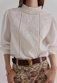 Witte geborduurde blouse met hoge kanten kraag, voorzien van verticale uitsparingen en luchtige mouwen, gecombineerd met een patroonbroek en een riem met gouden accenten.