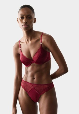 Mujer de piel oscura con sujetador de encaje rojo y ropa interior a juego, de pie frente a un fondo claro y sencillo.
