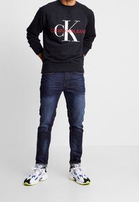 Uomo che indossa una felpa nera di Calvin Klein Jeans, jeans blu scuro e sneaker bianche con accenti blu e verde neon, in piedi con le mani dietro la schiena.
