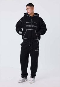 Sudadera negra con costuras en contraste y bolsillo frontal; combinada con joggers negros con franjas laterales y logotipos. Completado con zapatillas blancas.