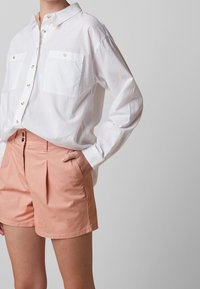 Camisa blanca abotonada con dos bolsillos en el pecho, combinada con pantalones cortos de color durazno. La camisa tiene un cuello clásico y un ajuste relajado.